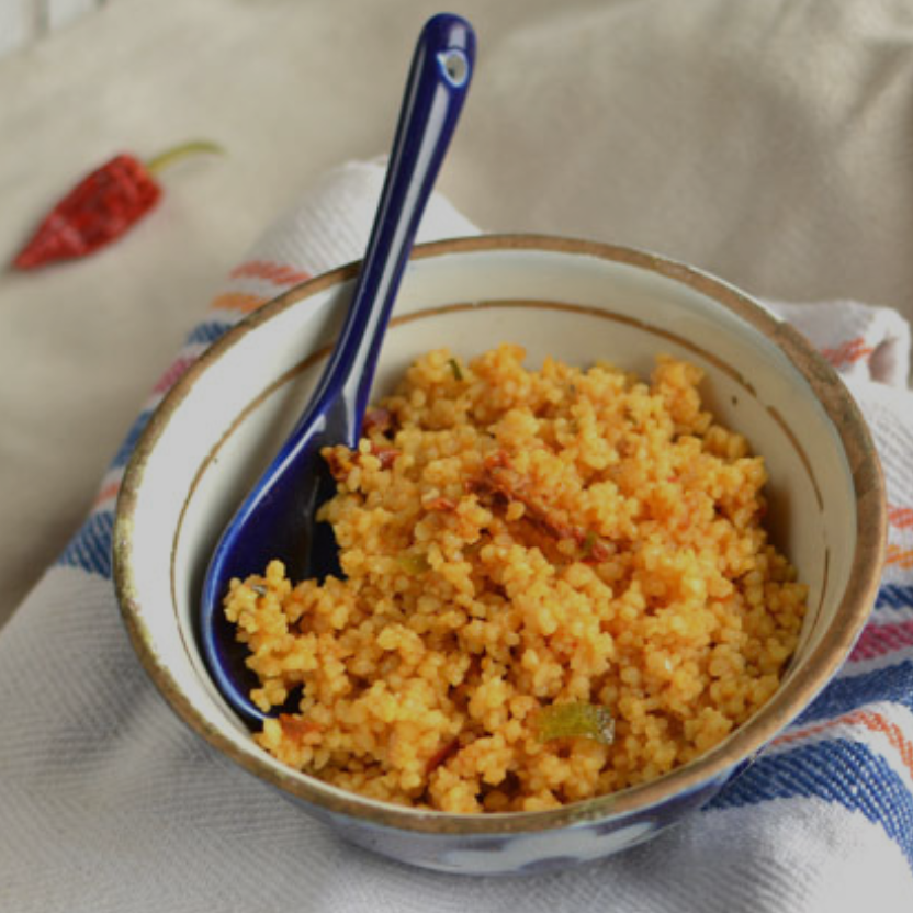 Couscous alla Marocchina in ciotola bianca e blu con cucchiaio blu, su strofinaccio a righe rosse e blu, peperoncino rosso sullo sfondo.