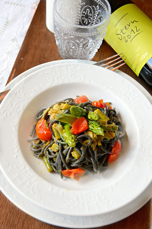 Spaghetti neri con pomodorini rossi e asparagi verdi in piatto bianco decorato, bicchiere e bottiglia di vino Stern 2012.