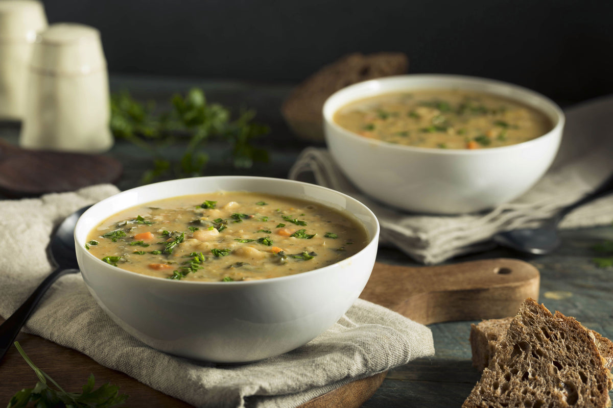 Due ciotole bianche di zuppa di fagioli pugliese con carote, prezzemolo e pane integrale su tagliere di legno.